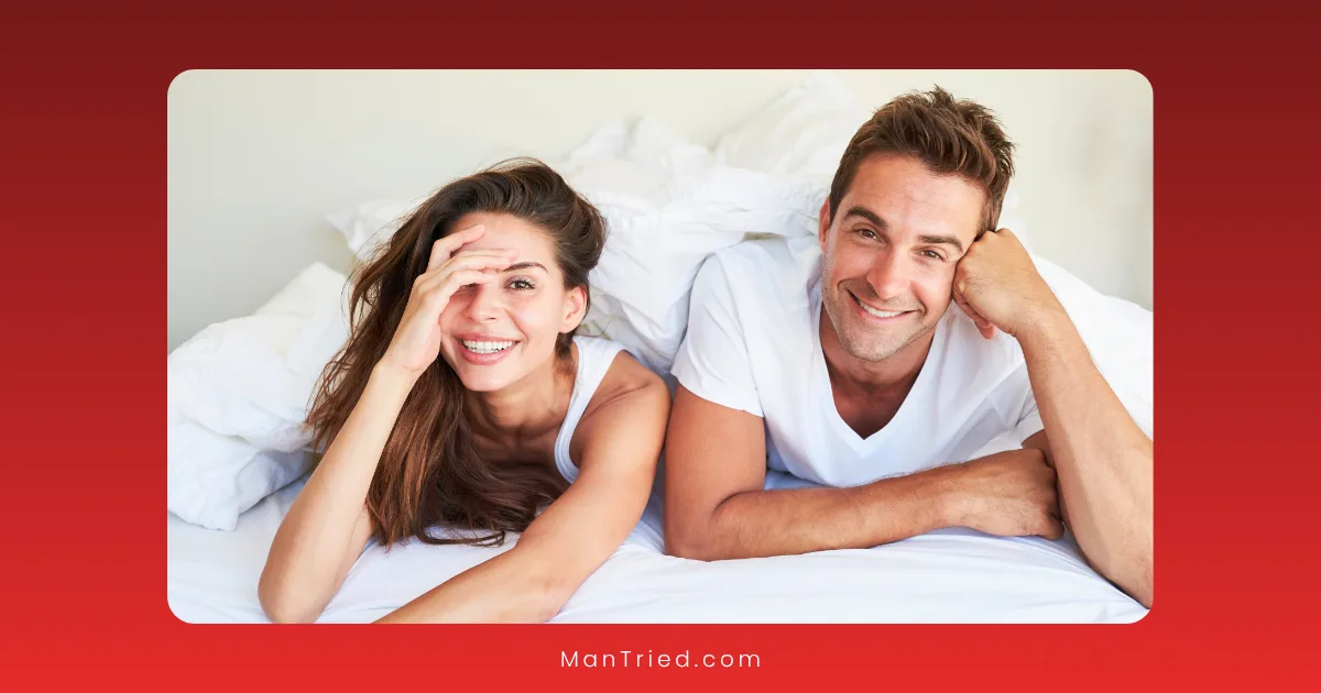 A smiling woman and man lie on a bed under white blankets, looking at the camera—sharing a moment that could be part of many PE recovery stories. The woman playfully covers part of her face while both look relaxed and happy against the bright white background.