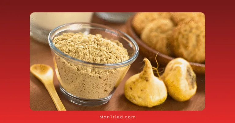 A clear glass bowl filled with light brown maca powder sits on a wooden surface next to two whole maca root and a small wooden spoon. Blurred bread is visible in the background.
