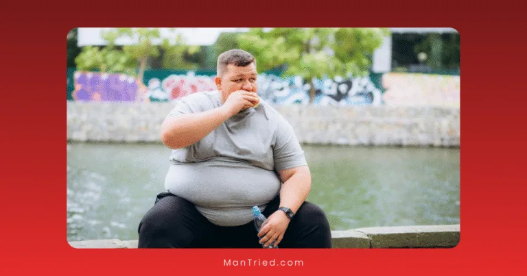 A man in a gray t-shirt and black pants, potentially mindful of obesity and testosterone levels, sits outdoors by a river eating a sandwich and holding a bottle of water, with trees and graffiti in the background.