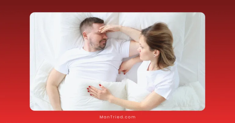 A woman and her partner dealing with delayed ejaculation lie in bed facing each other. The woman gently touches his forehead as if checking for a fever. Both wear white shirts under a white blanket, set against a white and red gradient background.