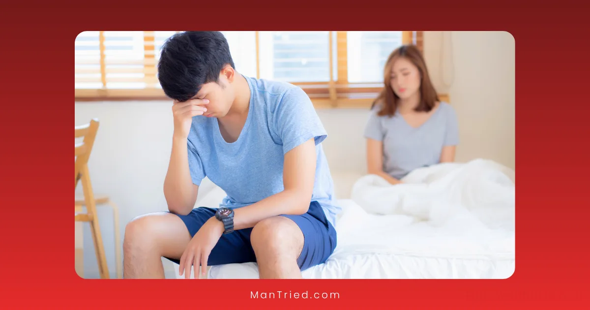 A man sits on the edge of a bed with his head in his hand, looking stressed—possibly due to premature ejaculation or performance anxiety—while a woman sits behind him, watching with concern. The room is softly lit with natural light.