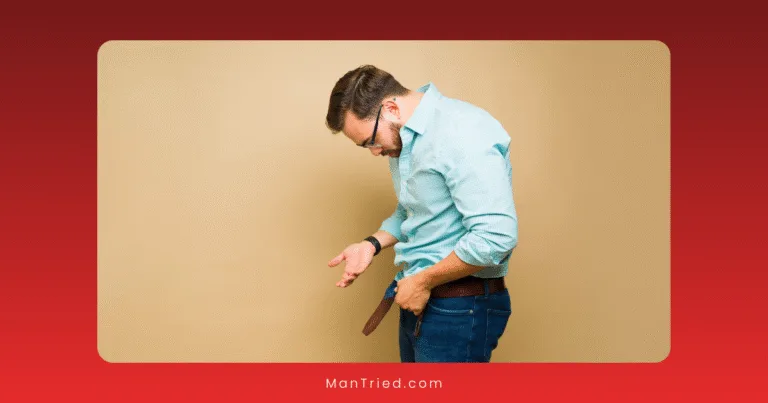 A man in a blue shirt and jeans looks down while adjusting his open belt against a plain beige background, hinting at concerns about the root causes of erectile dysfunction.