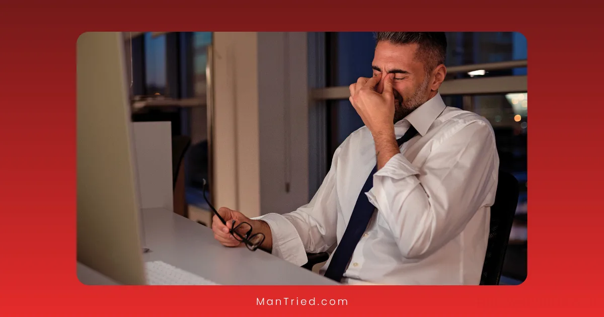 A man in a white shirt and tie sits at a desk, holding his glasses and rubbing the bridge of his nose, appearing stressed or tired—possibly due to the effects of shift work on his sleep and testosterone levels—while looking at a computer screen.