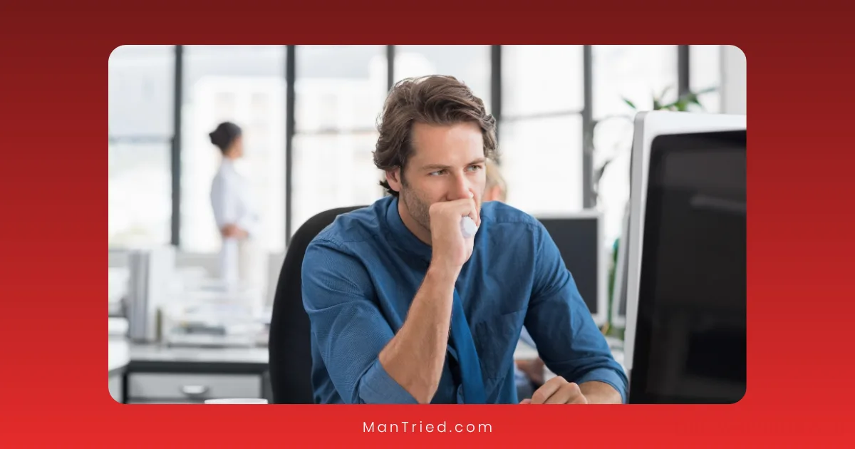 A man in a blue shirt sits at a desk in an office, looking thoughtfully at a computer monitor with his hand covering his mouth, possibly reflecting on stress and its impact on male libido. Two people are blurred in the background.