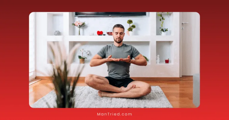 A man sits cross-legged on a rug in a living room, meditating with his eyes closed and hands raised—embracing stress-reduction techniques. The room features wooden floors, plants, and shelves with decorative items in the background.