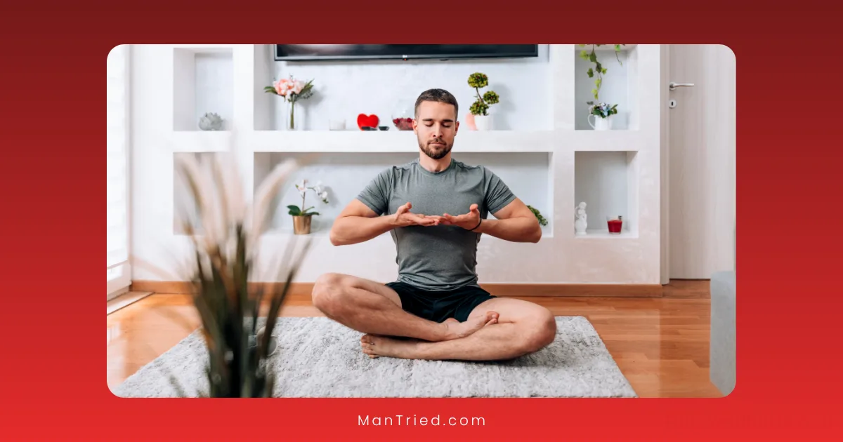 A man sits cross-legged on a rug in a living room, meditating with his eyes closed and hands raised—embracing stress-reduction techniques. The room features wooden floors, plants, and shelves with decorative items in the background.