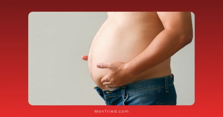 A person with a bare, protruding belly—likely due to visceral fat—stands sideways in unbuttoned blue jeans, holding their stomach with both hands against a plain background, highlighting potential links to issues like erectile dysfunction.