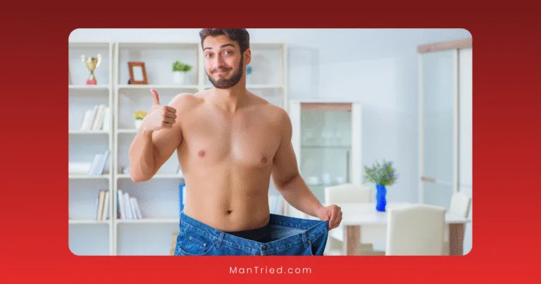 A shirtless man stands indoors, smiling and giving a thumbs-up while holding out the waistband of oversized jeans, proudly showing he’s lost 10% of his body weight. Shelves and a table are visible in the background.
