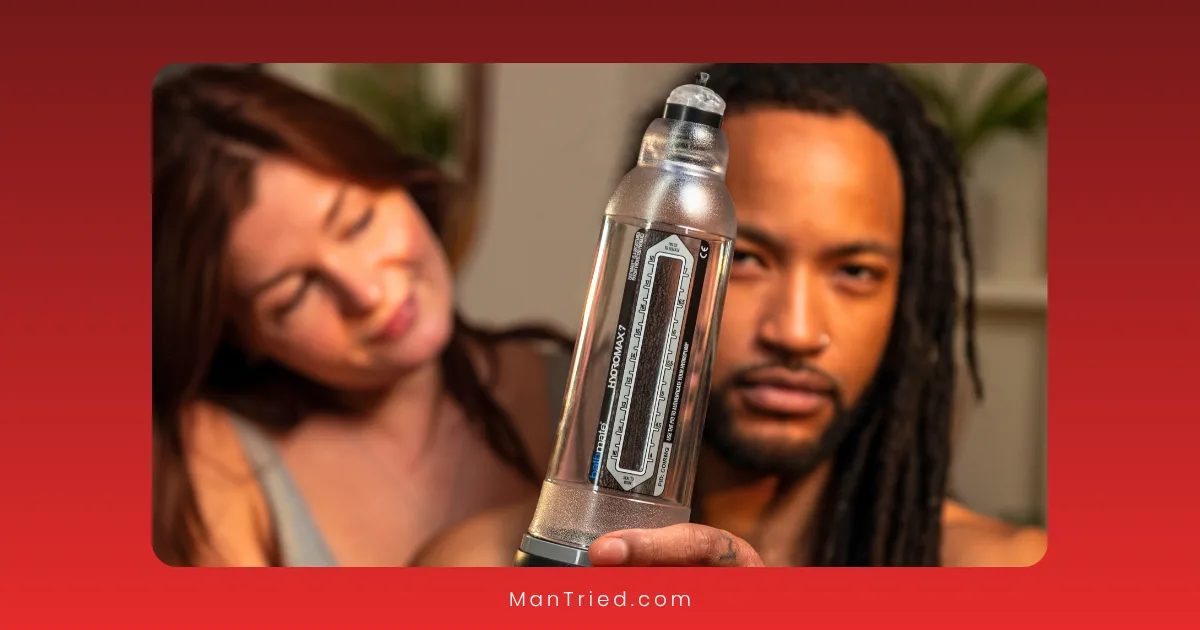 A man with long hair holds a clear cylindrical assistive device for sexual function toward the camera, while a woman with brown hair sits in the background, smiling and watching him. The background is softly blurred.