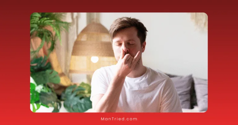 A man sits indoors wearing a white shirt, practicing alternate nostril breathing techniques with one hand on his nose. Plants and a lamp are in the background.