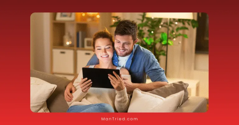 A smiling couple sits on a couch at home, enjoying digital communication about sex as they look at a tablet together. The room is warmly lit, with a plant and shelves in the background.