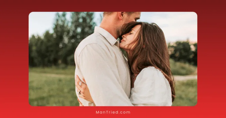 A couple shares a moment of emotional intimacy outdoors; the woman closes her eyes and leans into the man, who gently rests his chin on her forehead. They stand in a grassy field with trees behind them, both dressed in light-colored clothing.