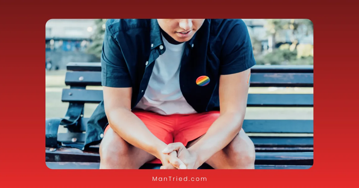A person in a black shirt with a rainbow pride pin, reflecting on minority stress and sexual function, sits on a bench leaning forward, hands clasped. The outdoor background is blurred.