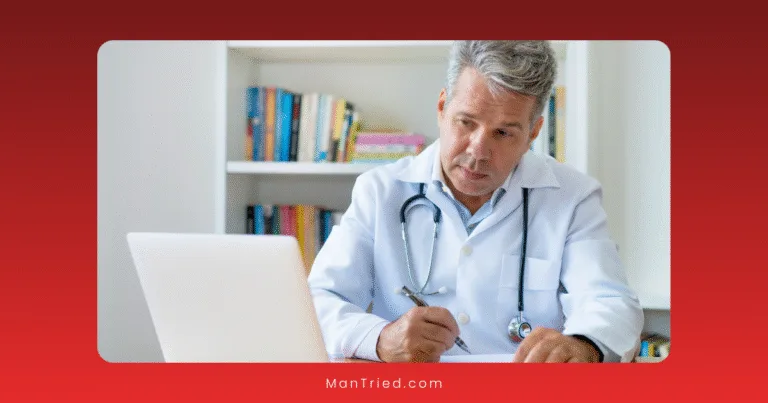 A middle-aged male oncologist with gray hair, wearing a white coat and stethoscope, writes notes while looking at a laptop in an office with a bookshelf in the background.