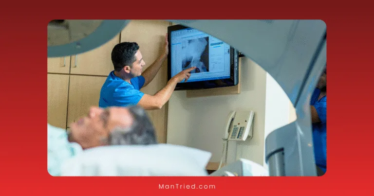 A medical professional in blue scrubs points at an X-ray image on a screen while discussing penile rehabilitation after radiation, as a patient lies on a hospital bed in the clinical room foreground.