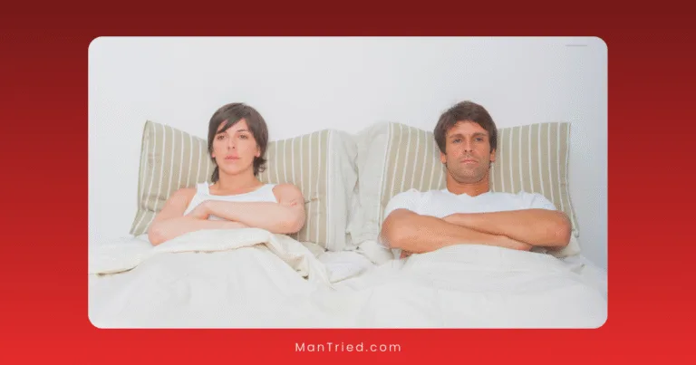 A man and woman sit up in bed with arms crossed, looking serious and distant—possibly reflecting on performance anxiety vs ED—under white bedding with striped pillows against a plain white wall.