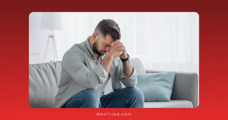 A man sits on a couch with his elbows on his knees, head bowed, and hands clasped in front of his face, appearing stressed or deep in thought under performance pressure.