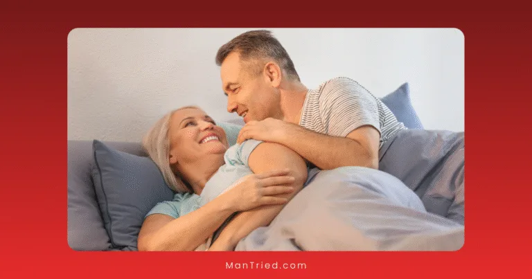 A smiling mature couple lies in bed together, facing each other and sharing an affectionate moment under a light blue blanket, celebrating intimacy and sex after 50. The background features a soft, white wall.