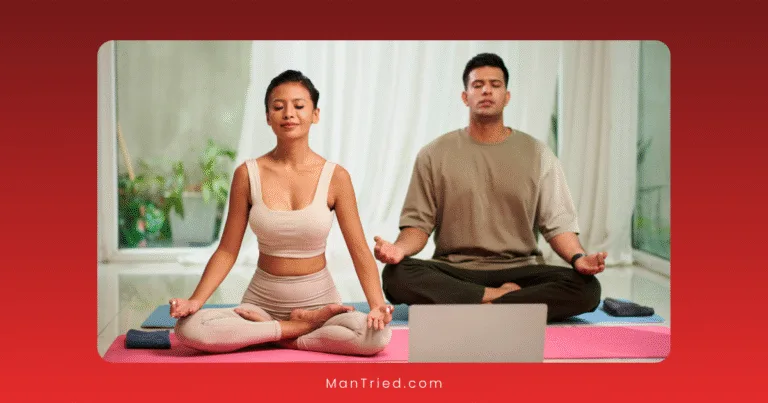 A woman and a man sit cross-legged on yoga mats indoors, meditating with closed eyes and hands on their knees, practicing tantric breathing as they face a laptop; the softly lit background adds to the calming atmosphere.