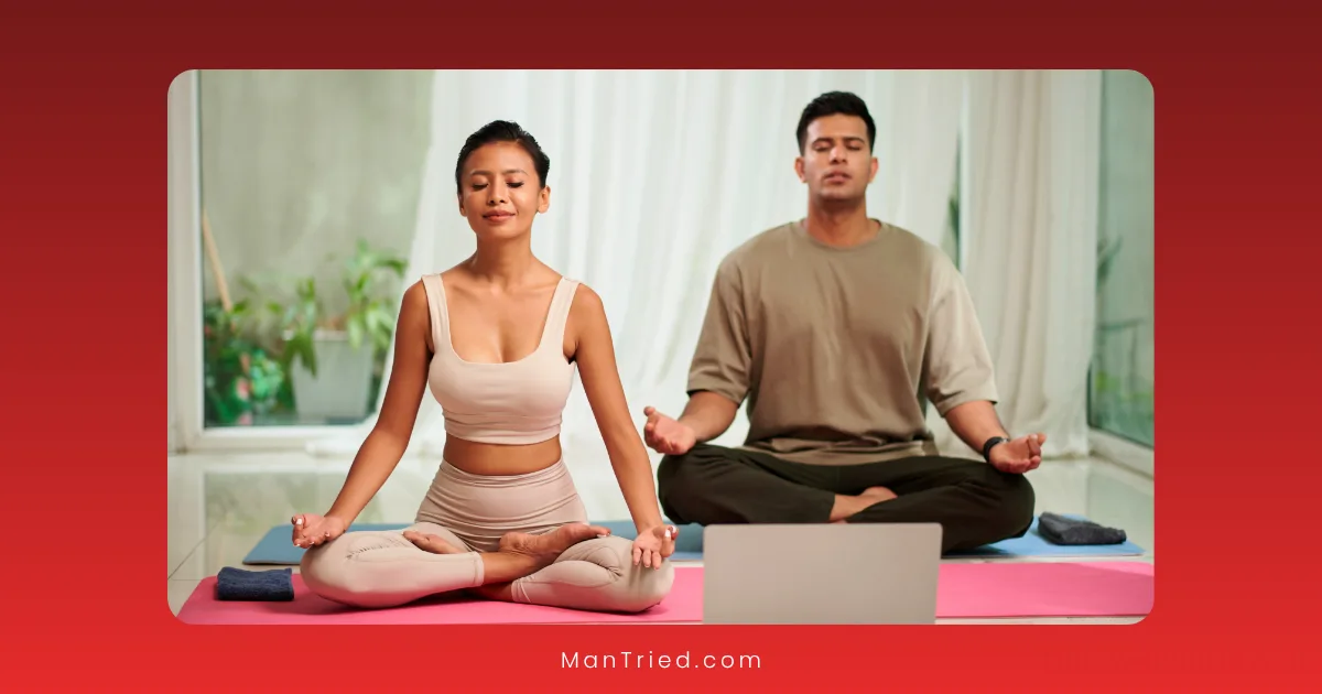 A woman and a man sit cross-legged on yoga mats indoors, meditating with closed eyes and hands on their knees, practicing tantric breathing as they face a laptop; the softly lit background adds to the calming atmosphere.
