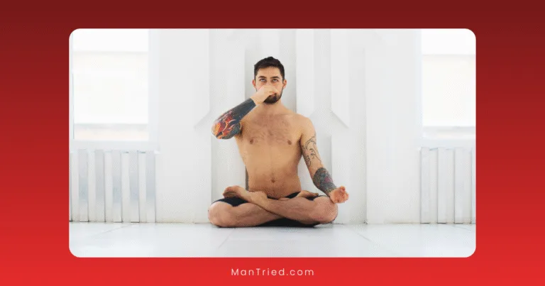 A shirtless man with tattoos sits cross-legged on the floor in a bright room, practicing a tantric breathing exercise by closing one nostril with his hand.