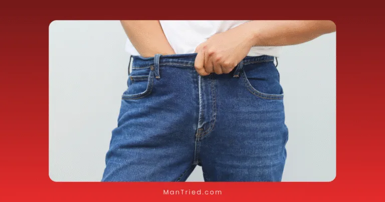 A person wearing a white shirt uses the edge method to pull up the waistband of blue jeans with one hand against a plain background.
